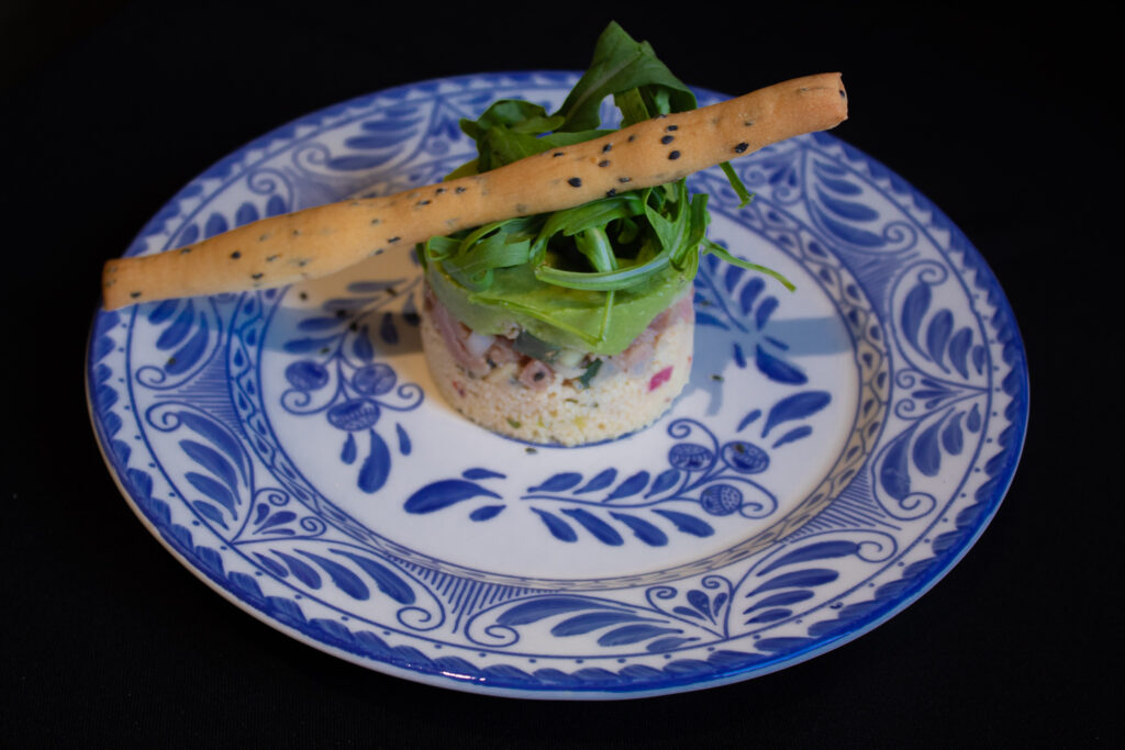 Montaje de entrada individual con couscous, aguacate y arúgula, decorado con grissini de ajonjolí, servido en vajilla artesanal mexicana azul.